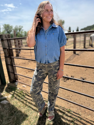 Woman in a blue shirt and camouflage pants standing in an outdoor setting with a fence and trees in the background.