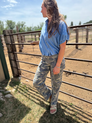 Woman in blue shirt and camouflage pants standing near a wooden fence with a rural landscape in the background.