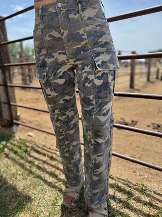 Camouflage pants worn by a person standing in an outdoor setting with a metal fence and grassy area.