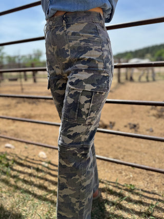 Camouflage pants worn by a person standing in an outdoor setting with a fence and grass in the background.