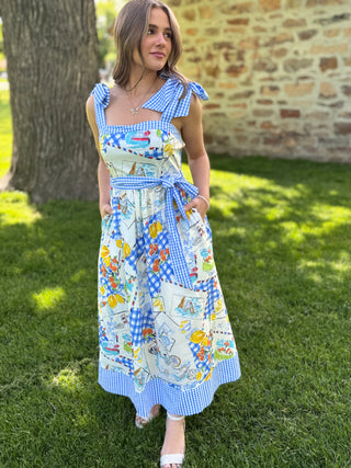 Woman wearing a colorful patchwork dress standing outdoors on grass with a brick wall in the background.