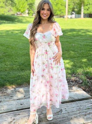 Woman in a floral dress standing on wooden planks with grass and trees in the background