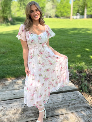 Woman in a floral dress standing on a wooden platform with grass and trees in the background