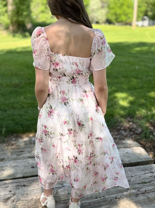 Woman wearing a floral dress standing on a wooden platform with green grass and trees in the background