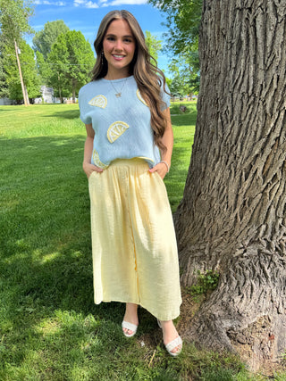 Woman in a light blue top with yellow patterns and a yellow skirt standing next to a tree in a park.