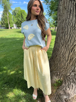 Woman in a light blue sleeveless top with yellow patterns and a yellow skirt standing next to a tree in a park.