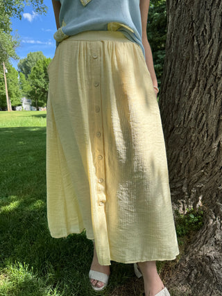 Person wearing a light yellow skirt with buttons standing next to a tree outdoors.