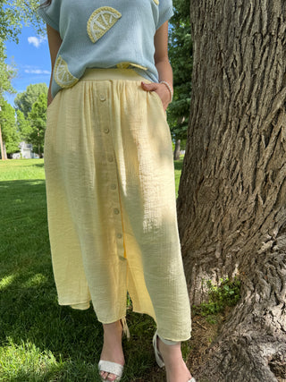 Person wearing a light blue shirt with lemon design and yellow pants standing next to a tree in a park.