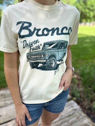Person wearing a t-shirt with 'Bronco Driven Faith' graphic outdoors.