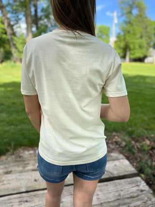 Person wearing a light-colored t-shirt and denim shorts standing on a wooden deck with a grassy area and trees in the background.