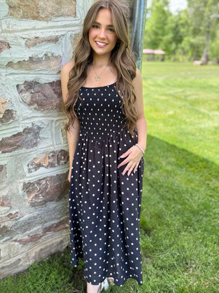 Woman in a black polka dot dress standing outdoors near a stone wall.