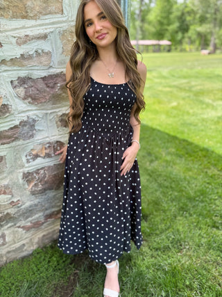Woman in a black polka dot dress standing outdoors near a stone wall.