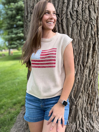 Woman wearing a sleeveless top with an American flag design, standing next to a tree in a park.