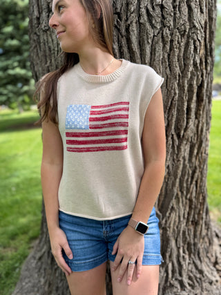 Woman wearing a sleeveless sweater with an American flag design, standing next to a tree outdoors.