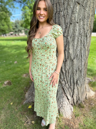 Woman in a green floral dress standing next to a tree in a park