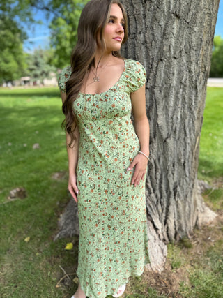 Woman in a green floral dress standing next to a tree in a park