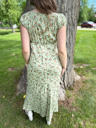 Person wearing a green floral dress standing next to a tree in a park.