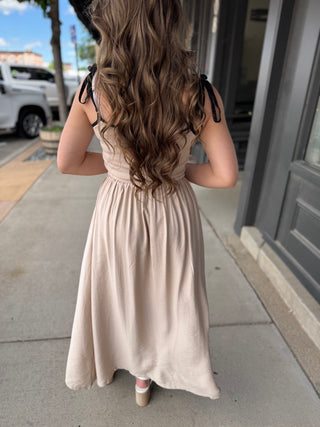 Woman in a beige dress standing on a sidewalk with a building and parked cars in the background.