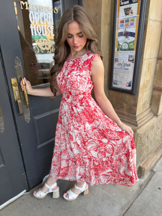 Woman in a red and white floral dress standing outside a building with a door and window.