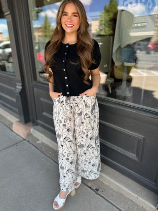 Woman wearing a black top and white floral pants standing on a sidewalk.
