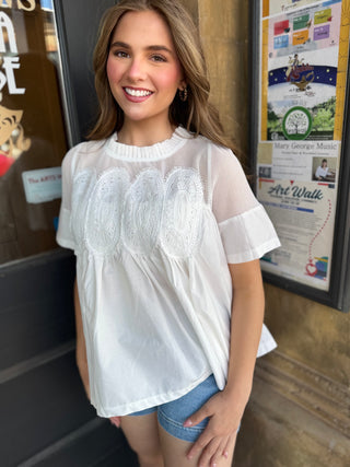 A woman wearing a white short-sleeve blouse with three large embroidered oval motifs on the front, paired with denim shorts, standing in front of a storefront.