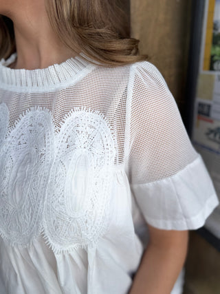 A close-up of a person wearing a white blouse with a sheer mesh yoke, embroidered lace detailing across the chest, and short flared sleeves.