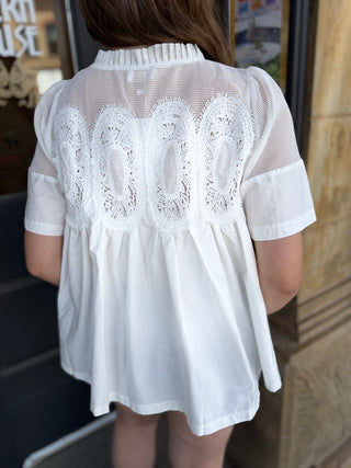 A woman viewed from behind wearing a white short-sleeve blouse with sheer mesh sleeves and a decorative white lace yoke across the upper back.