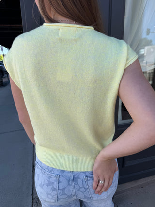 Person wearing a light green sleeveless sweater and blue jeans, standing outdoors.