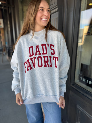 Woman wearing a gray hoodie with 'DAD'S FAVORIT' in red text, standing outside a building.
