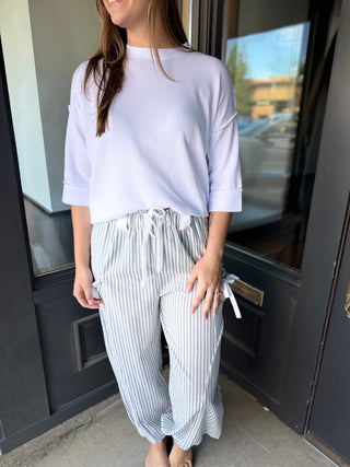 Woman wearing a light purple top and striped pants standing in front of a door.