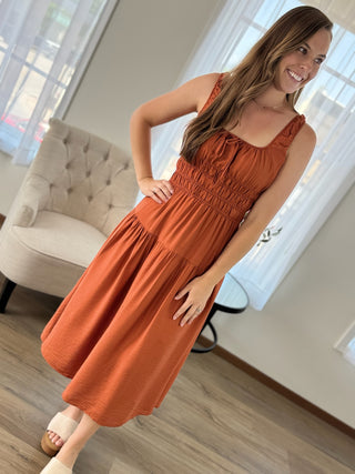 A woman modeling a rust-orange sleeveless midi dress with a smocked bodice and tiered skirt, standing in a brightly lit room with a tufted armchair and windows in the background.