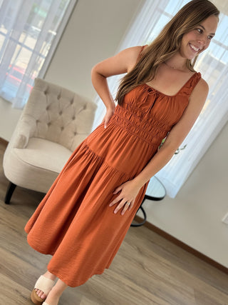 A woman wearing a sleeveless rust-orange tiered midi dress with a smocked waist, standing in a brightly lit room with a tufted armchair and windows in the background.