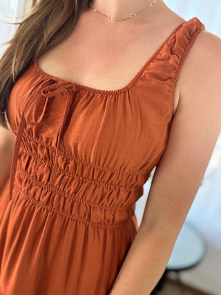 Close-up of a woman wearing a rust-orange sleeveless dress with a smocked bodice, ruffled straps, and a tie detail at the neckline.