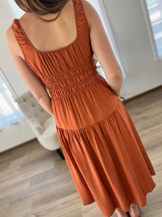 A woman wearing a terracotta orange sleeveless dress, shown from the back in a brightly lit indoor room with light wood floors and a white tufted chair.