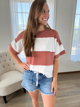 A woman wearing a short-sleeve striped t-shirt with dusty rose and white horizontal bands, paired with denim shorts, standing in a brightly lit room.