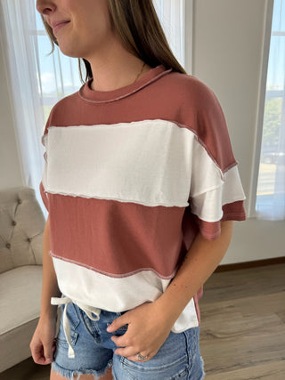 A woman wearing a short-sleeve color-blocked t-shirt with horizontal stripes of dusty rose and off-white, paired with denim shorts in a brightly lit room.