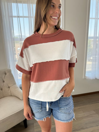 A woman wearing a short-sleeve color-blocked t-shirt with horizontal stripes in terracotta and off-white, paired with denim shorts, standing in a brightly lit room.