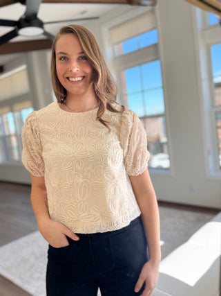 A woman smiling and wearing a beige embroidered top with puff sleeves.