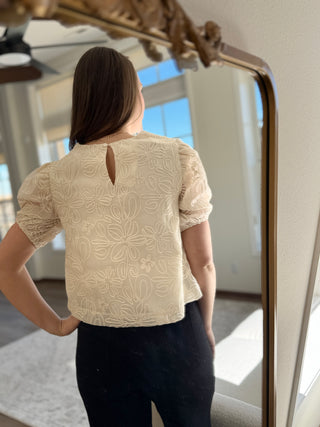 A woman smiling and wearing a beige embroidered top with puff sleeves.