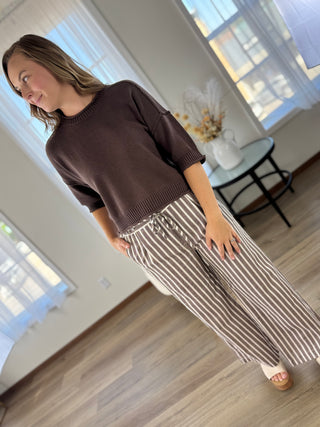 A woman modeling a dark brown short-sleeve knit sweater and brown-and-white vertically striped wide-leg pants in a bright room with wood floors and large windows.