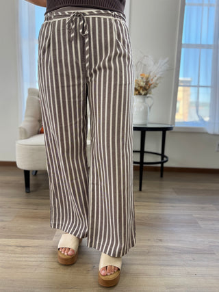 A person wearing wide-leg cropped pants with vertical brown and beige stripes, paired with cream platform sandals in a bright indoor room with wood flooring and a window.