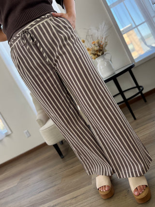 A person wearing brown and beige vertically striped wide-leg pants, paired with a dark brown ribbed top and beige platform sandals, standing in a modern room with wood floors and a window.