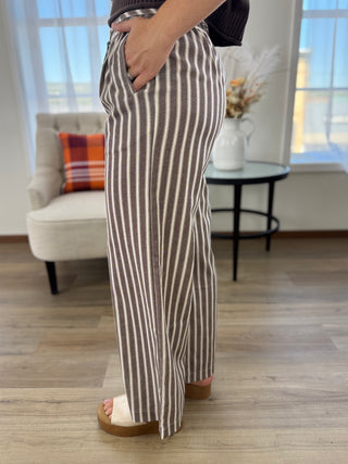 A person wearing high-waisted, wide-leg trousers with vertical brown and beige stripes, standing in a bright living room with wood floors and a window.