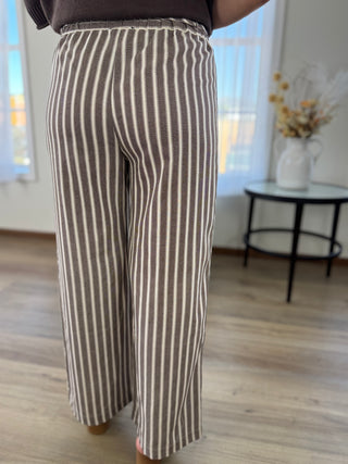 A person wearing loose, wide-leg cropped pants with vertical brown and off-white stripes, standing in a bright indoor room with wood flooring and a small table in the background.