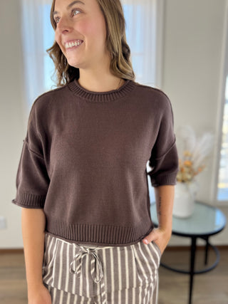 A woman wearing a cropped, short-sleeve knit sweater in a muted brown color, paired with beige and white striped drawstring pants, standing in a brightly lit indoor room.