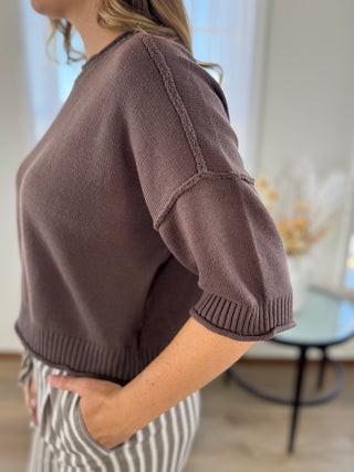 A woman wearing a cropped, short-sleeve knit sweater in a muted taupe-brown color, paired with gray and white striped pants, shown in a softly lit indoor setting.
