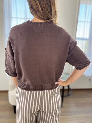 A woman shown from the back wearing a short-sleeve knit sweater in a muted brown color, paired with beige and white vertically striped pants, in a brightly lit room.