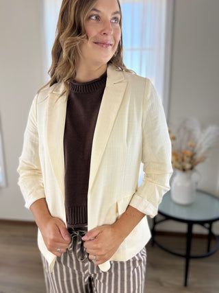 A woman wearing a cream-colored linen-textured blazer over a dark brown ribbed sweater and brown-and-white vertically striped pants, standing in a brightly lit indoor room.