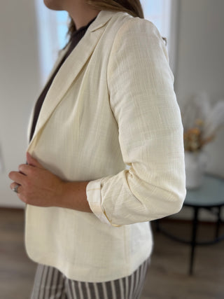 A woman wearing a light cream-colored textured linen-blend blazer over a dark top and striped trousers, shown in a side profile within a softly lit indoor room.
