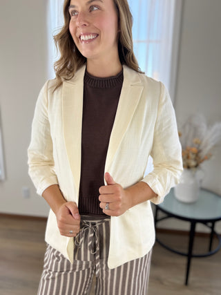 A woman wearing a cream-colored linen-textured blazer over a dark brown ribbed knit top and brown-and-beige vertically striped pants, standing in a brightly lit indoor room.